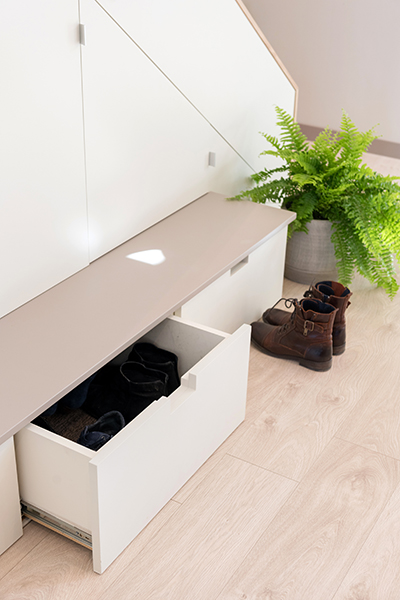 Rangement sous escalier avec banc avec tiroirs pour chaussures et placard. L'ensemble est blanc cassé et le plateau du banc taupe. Le parquet est beige clair. Une fougère dans un pot gris est au bout du banc.