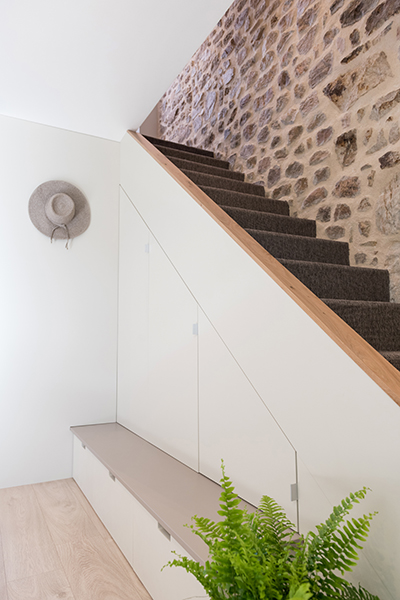 Aménagement sous les escaliers avec partie basse en banc avec tiroirs et partie haute placard. L'ensemble est blanc cassé. Les marches des escaliers sont en sisal brun, la rampe en chêne et le mur de la montée des escaliers en pierres apparentes.