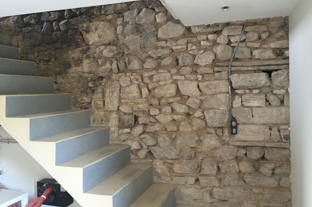 Maison en pierre en cours de rénovation. Vue sur escalier droit en béton devant un mur en pierres apparentes non jointées.