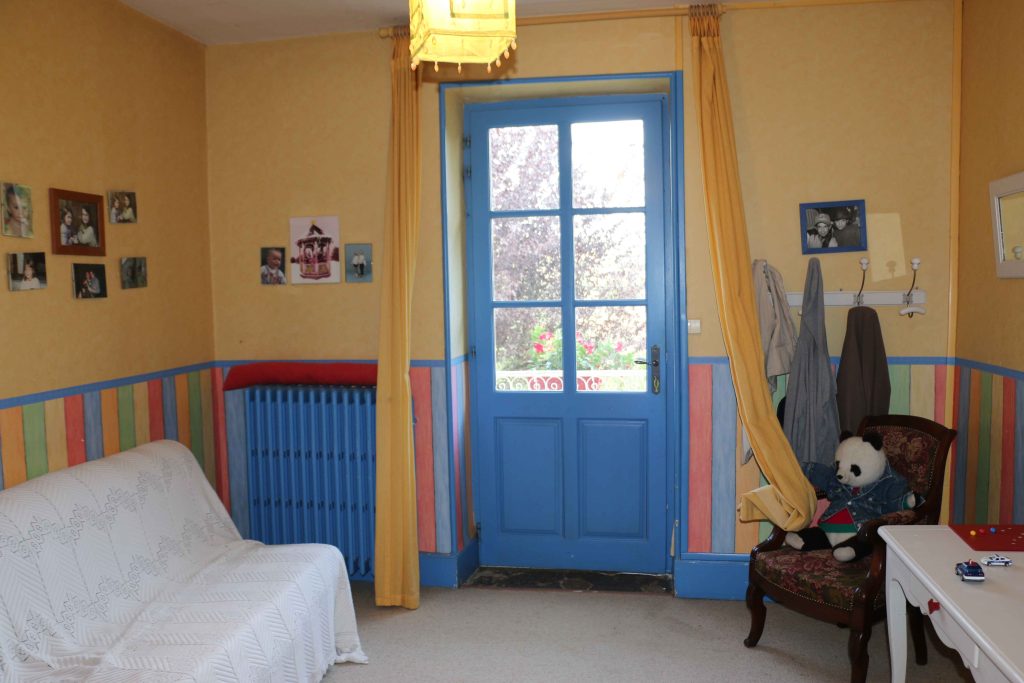 Petit salon d'une maison ancienne avec une porte à moitié vitrée bleue, Murs en papier-peint jaune avec sous-bassement rayures larges multicolores. Au sol une moquette grise. A gauche un canapé clic-clac avec une couverture blanche par-dessus, à droite un fauteuil ancien et un petit bureau.