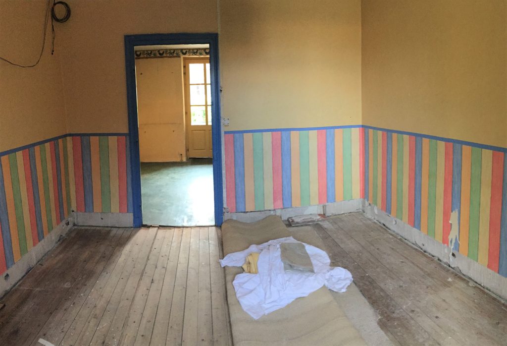 Pièce en cours de rénovation avec parquet ancien en pin. Mur en papier-peint jaune avec sous-bassement rayures larges multicolores. Cette pièce donne accès à une deuxième au sol vert.