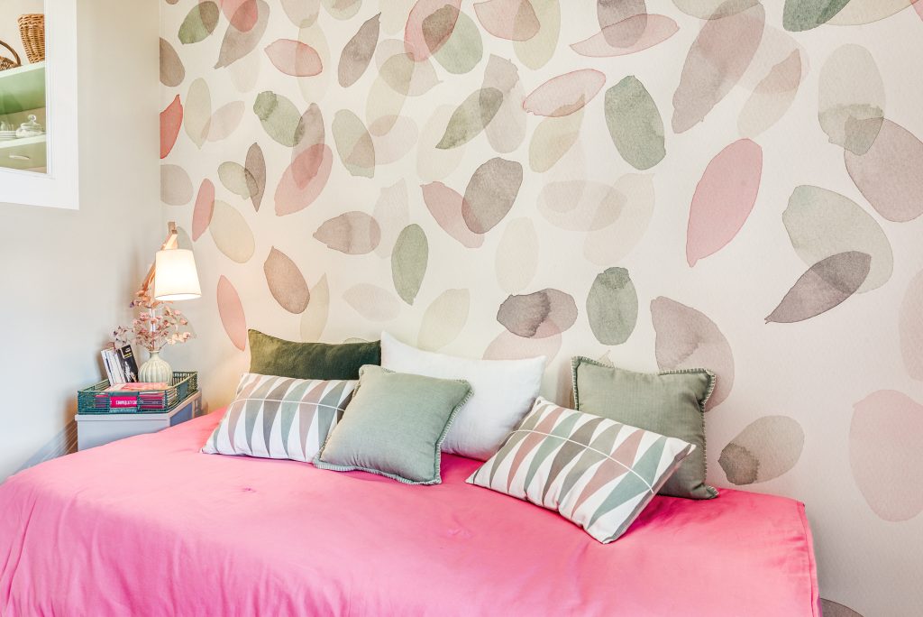 Lit utilisé comme méridienne avec drap rose et de nombreux coussins dans les tons verts pastel, rose et blanc. A gauche une table de chevet avec une lampe avec abat-jour blanc. Le mur est habillé d'un papier peint représentant une cascade de pétales en aquarelle dans les couleurs pastels rose, vert et taupe.