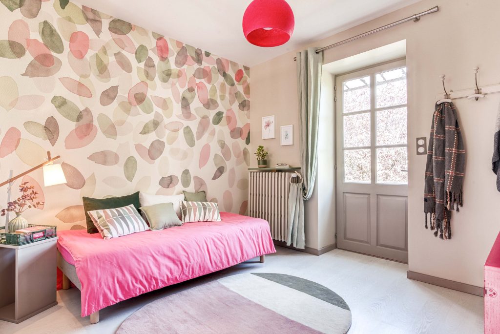 Petit salon cosy dans des teintes pastel de rose, vert et taupe. Lit utilisé comme méridienne avec drap rose et de nombreux coussins dans les tons verts pastel, rose et blanc. A gauche une table de chevet avec une lampe avec abat-jour blanc. Le mur est habillé d'un papier-peint représentant une cascade de pétales en aquarelle dans les couleurs pastels rose, vert et taupe. Sur le sol en parquet blanc, il y a un tapis ovale dans les mêmes teintes.