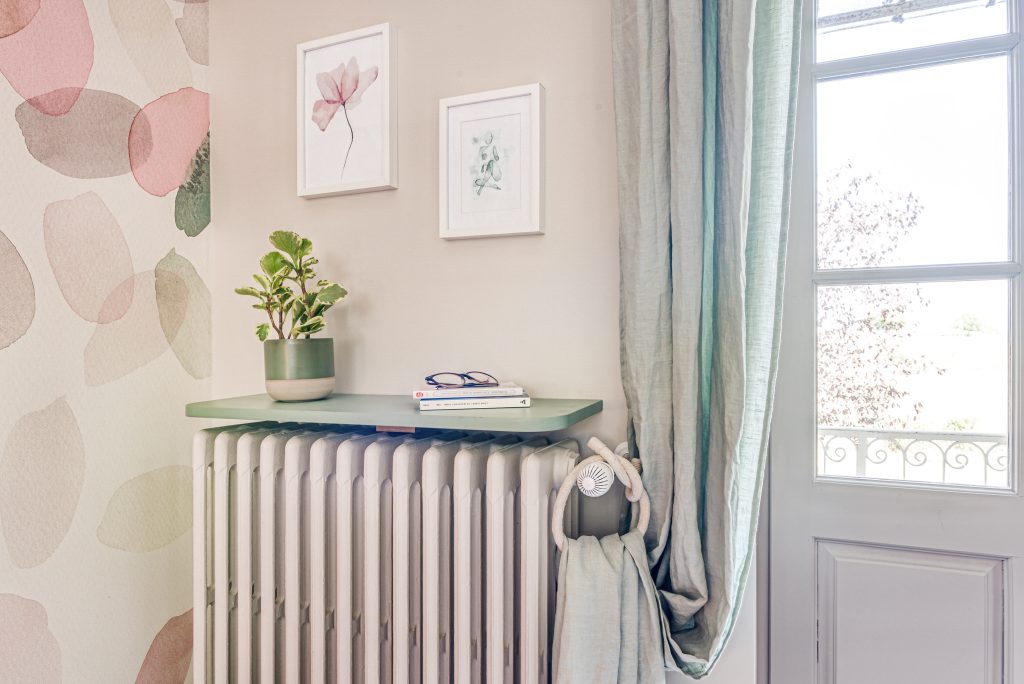 Zoom sur un radiateur en fonte peint en taupe comme les murs. Avec au-dessus une tablette verte tilleuil sur laquelle est disposée une petite plante en pot, des livres de poche et des lunettes. Sur le mur au-dessus, il y a des aquarelles de fleurs rose et vert pastel, qui vont avec le papier-peint du mur de gauche (pétales en aquarelle parsemées et superposées de couleur rose, taupe, vert pastel). Au droite, il y a une porte à moitié vitrée donnant sur le jardin, et un rideau vert d'eau.
