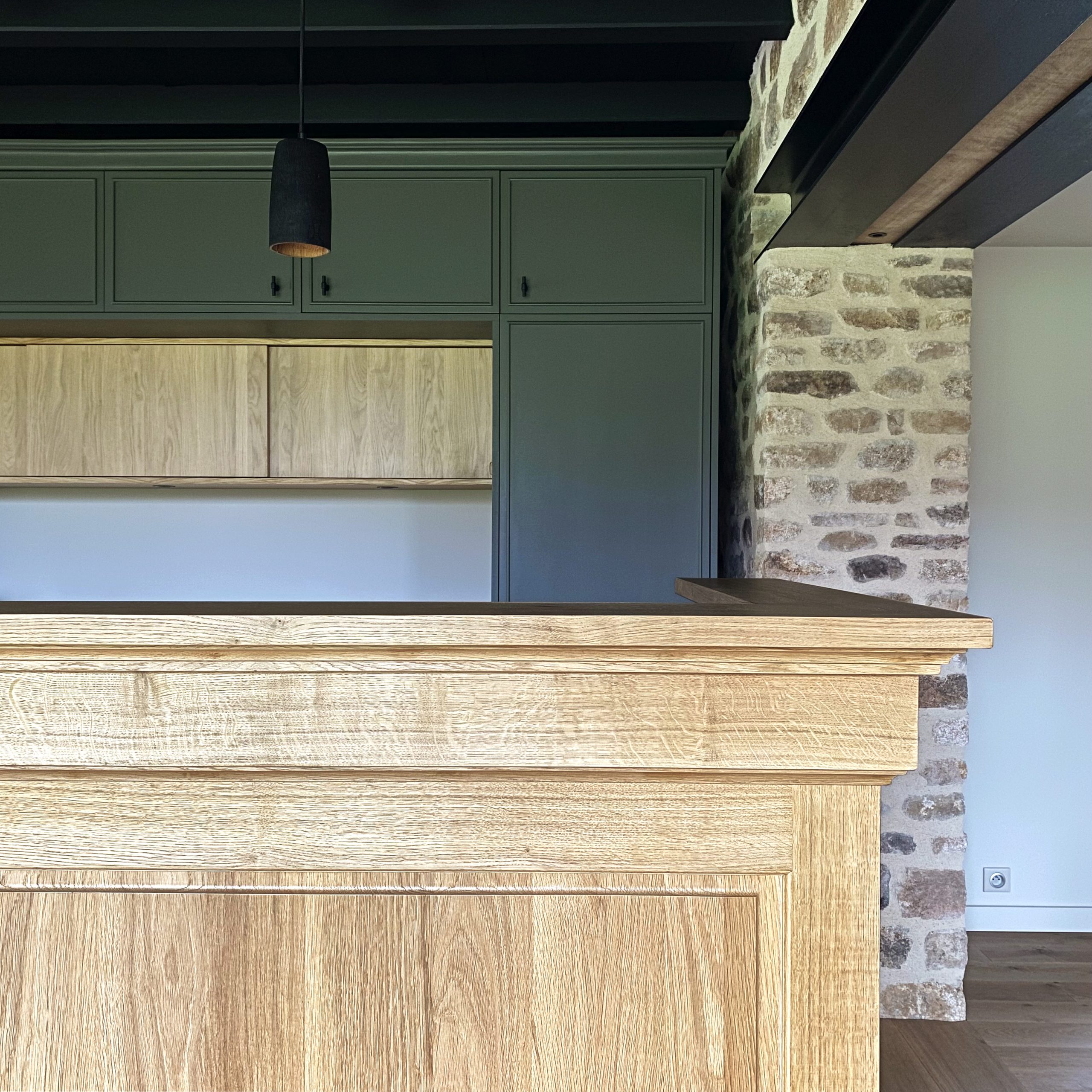 Cuisine sur mesure avec grand îlot en chêne massif et meubles verts profonds, murs en pierre apparente et plafond en poutres noires apparentes.