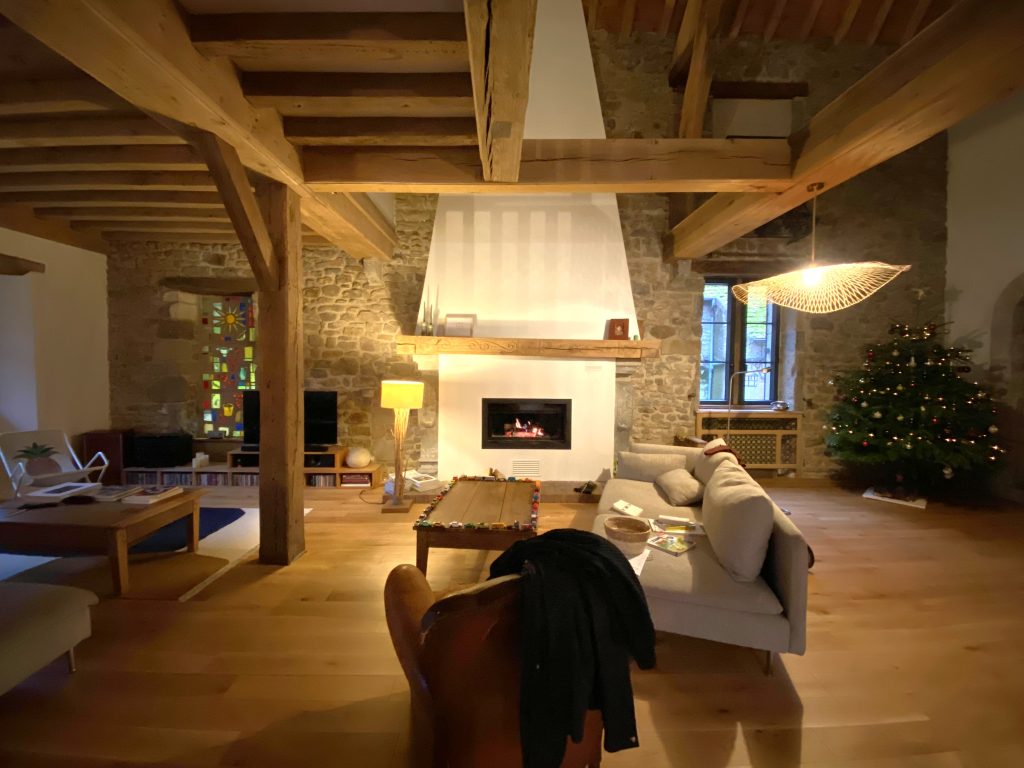 Coin détente devant grande cheminée ancienne renovée avec un insert. Plafond en poutre apparente et mur de la cheminée en pierres apparentes. Petit salon avec canapé gris au centre, sur la gauche espace TV et sur la droite un sapin de Noël.