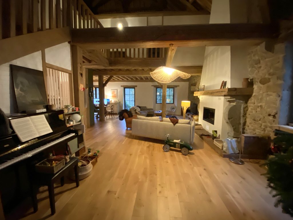 Coin musique avec piano droit noir sur la gauche, en arrière-plan un salon devant la cheminée en pierre avec un canapé gris et un fauteuil en cuir marron.