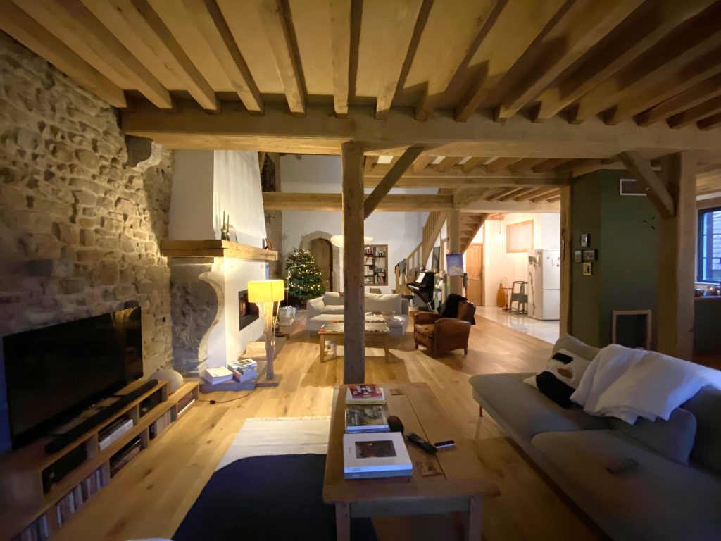 Salon TV cosy et sombre sous plafond bois avec poutres avec canapé gris, en arrière-plan un deuxième salon devant la cheminée en pierre avec un canapé gris et un fauteuil en cuir marron.