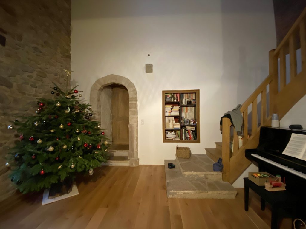 Coin d'un salon devant montée d'escalier en bois, à droite un piano droit noir et à gauche un sapin de Noël. Mur blanc et parquet chêne.
